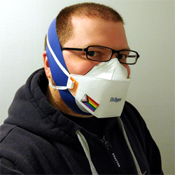 Photo of me: white guy in a navy blue hoodie with buzzed brown hair & beard wearing rectangular black glasses & a white Draeger 1950 trifold N95 adorned with a magnetic Pride flag pin. Under it is a blue, non-latex, exercise loop wrapped from below chin to crown of head, containing the beard for a better seal.